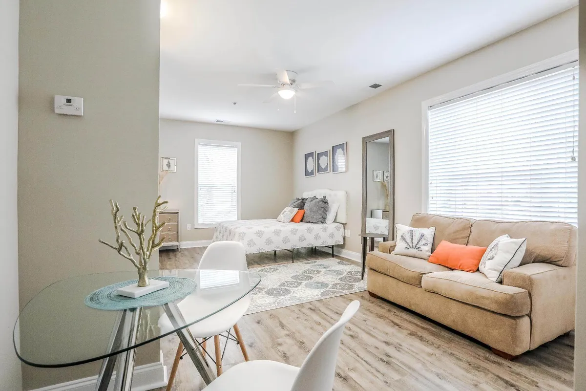 A view of the main living space featuring a glass dining table, a sofa with colorful cushions, and a bed with appealing patterned bedding. Framed artwork and decor elements add a contemporary touch, while ample natural light floods the area, creating an inviting and multi-functional environment.