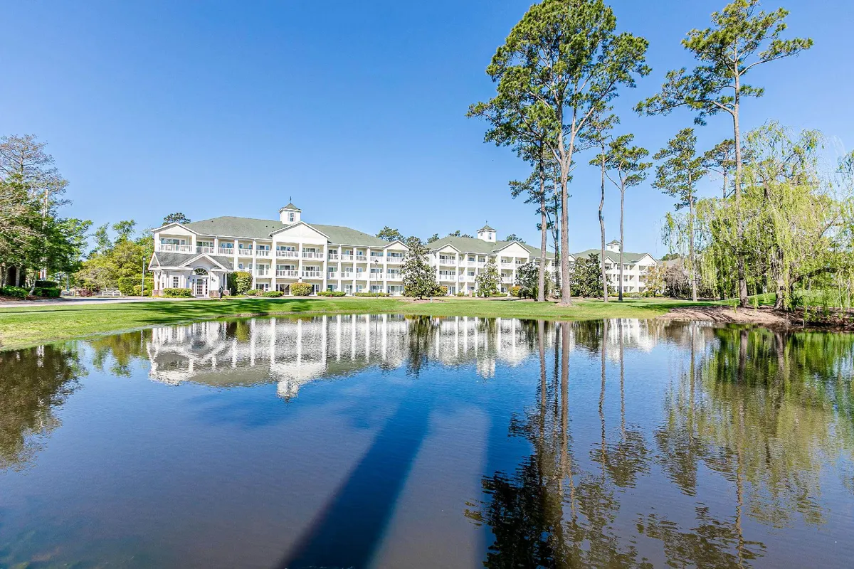 A peaceful exterior view of the Grande Villas condo complex, reflected in a tranquil pond and surrounded by tall trees under a clear blue sky. The lush green lawn invites visitors to relax outdoors or enjoy a sunset by the water.