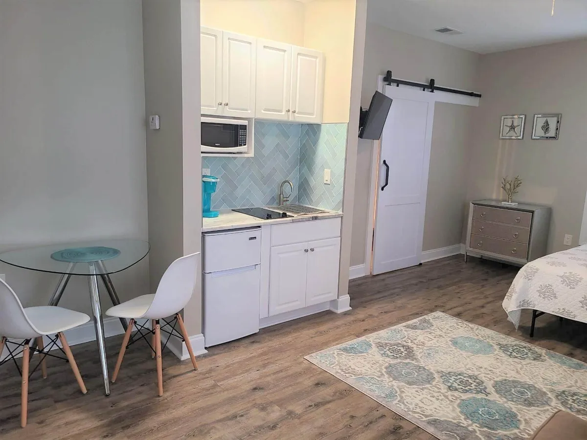 A bright, comfortable interior shows the kitchenette, dining table, and a decorative rug, all in one view. The image hints at the adjoining bed area, but the focus remains on the kitchen and dining space, emphasizing their cozy and modern design. Subtle details, like a coffee maker and toaster, reinforce the narrative of morning rituals and intimate meals.