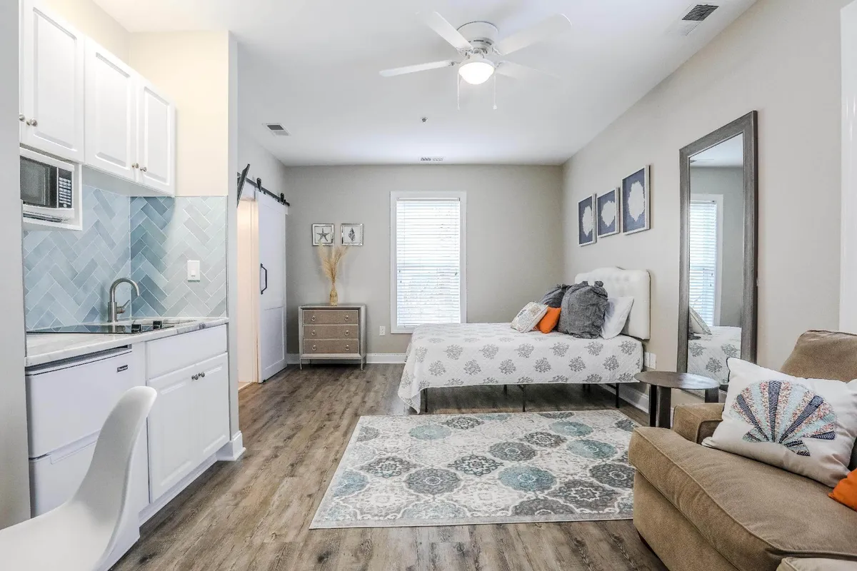 This wide-angle interior shot captures the studio’s main living area, offering a clear view of the bed with plush bedding, a cozy sofa, a stylish mirror, and the kitchenette. The decorative rug and warm decor accents create a modern yet inviting atmosphere. There are thoughtful touches such as reading materials on the table and a sense of openness that reflects the studio’s blend of comfort and functionality.