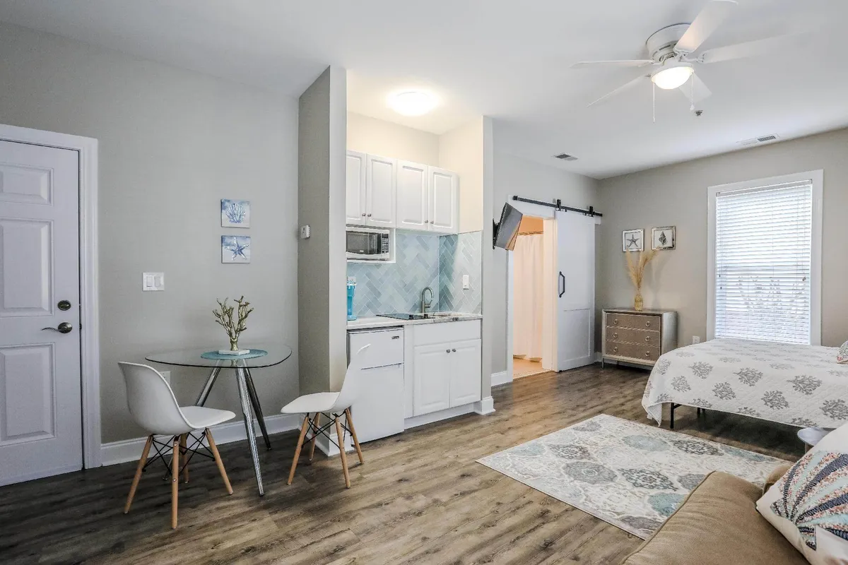 The image showcases a bright and inviting studio interior, featuring a cozy bed, a modern kitchenette perfect for preparing light meals, a small dining table ideal for intimate breakfasts, and stylish decorative wall art. The overall atmosphere is warm and relaxed, with natural light filtering in, emphasizing the condo's comfort and tranquility.