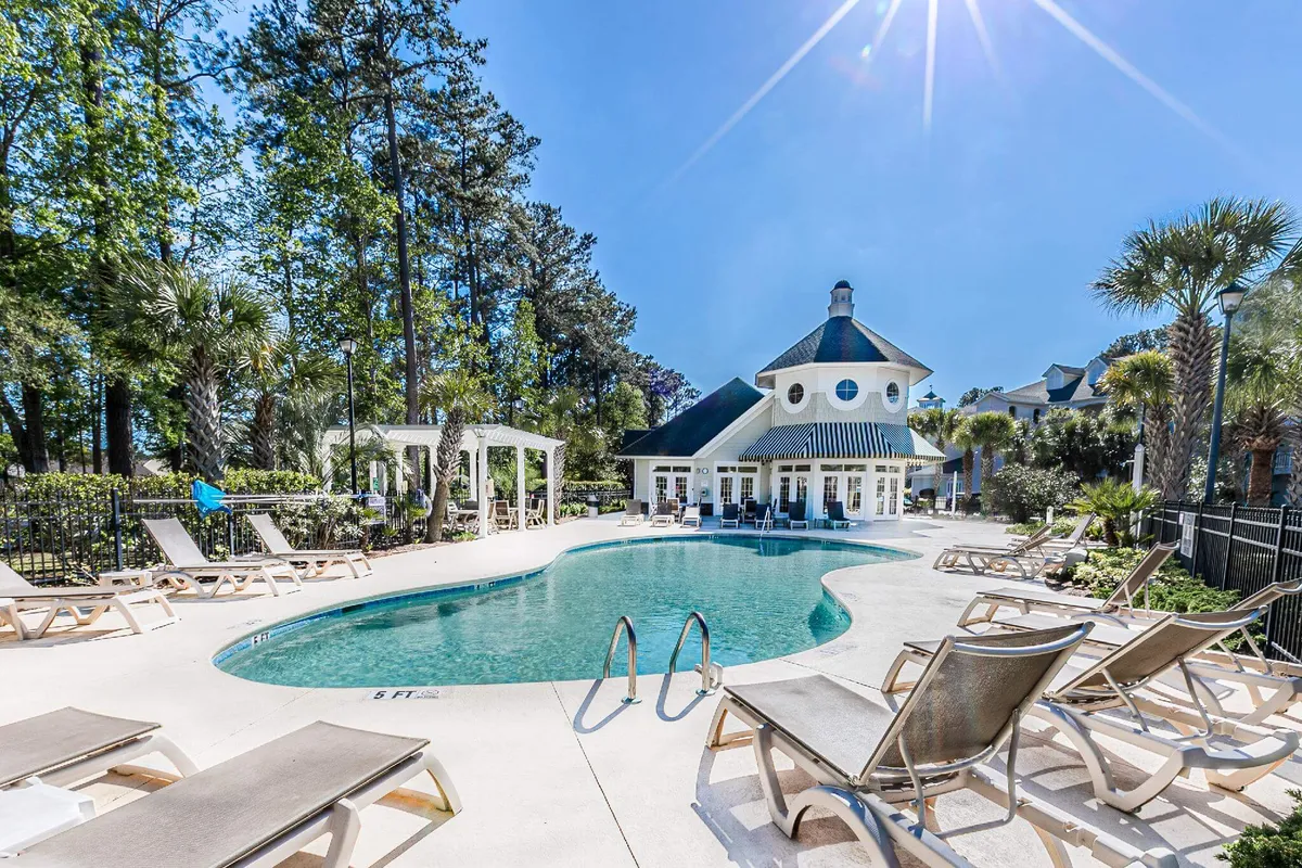 This image showcases the sparkling swimming pool surrounded by lounge chairs, with the elegant clubhouse in the background and lush palm trees dotting the landscape. The inviting pool area is vibrant, sunlit, and clearly designed for both relaxation and social gatherings.