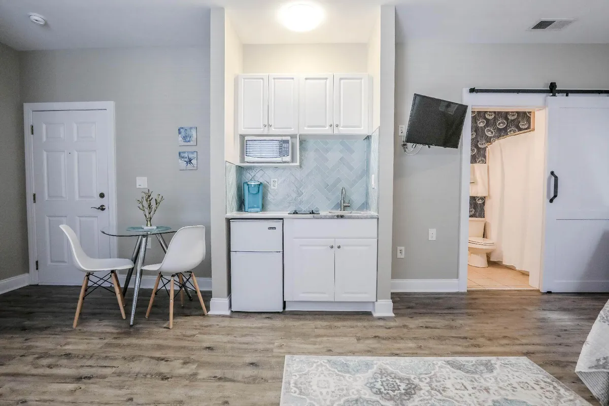 This image centers on the kitchenette and adjacent dining table within a comfortable, inviting interior. The well-equipped kitchen area is visible, with essential appliances and cooking basics on display. The proximity of the dining table highlights the space’s suitability for quick breakfasts, casual dinners, or enjoying a meal while watching TV. The overall ambiance is warm and practical.