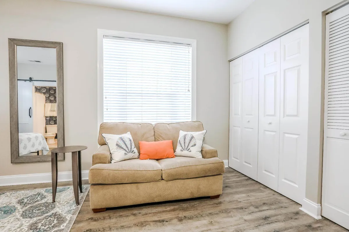 A comfortable living area featuring a plush sofa adorned with decorative pillows, a stylish side table, and a large mirror reflecting natural morning light. The setting is bright and welcoming, suggesting a spot for both relaxation and focused work.