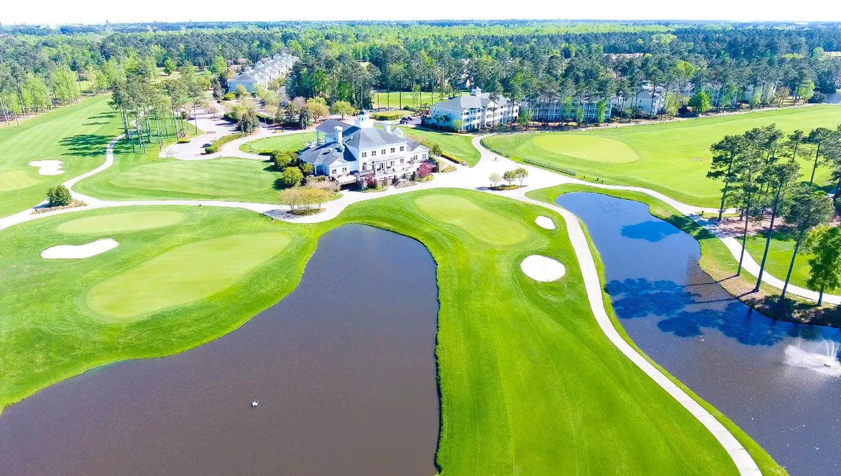 This image features a sweeping view of the World Tour Golf Course, with the inviting clubhouse and water features in the background. The setting captures the lush greens, tranquil water, and the central social hub of the clubhouse, where guests can gather after a round of golf.